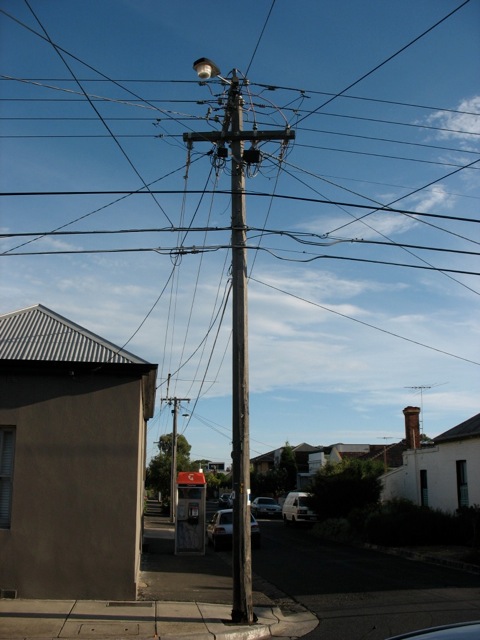A typical street scene in an inner suburb of Melbourne, showing an old wooden electricity pole (dangerously close to the curbing - a prime target to be hit by a truck) supporting two Pay TV cables, an electricity control cable, a street light and a multitude of electricity and pay TV lead-ins;